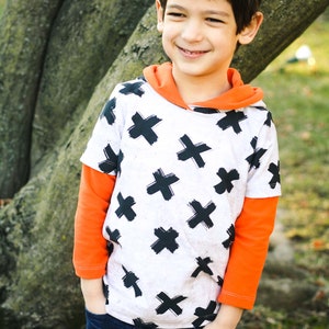 Può includere: Un bambino piccolo che indossa una felpa con cappuccio bianca e nera con maniche arancioni e jeans blu. La felpa ha un motivo di X nere su sfondo bianco.
