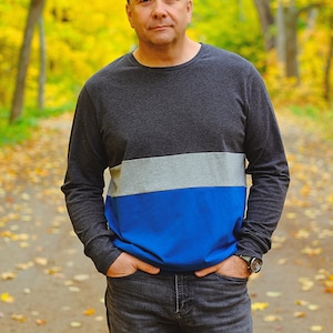 Puede incluir: Un hombre con una camisa de manga larga a rayas grises, azules y blancas con las manos en los bolsillos. Lleva pantalones vaqueros azules y un reloj en la muñeca izquierda. Está de pie en un bosque con hojas amarillas en el suelo.