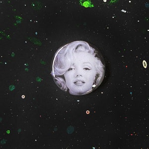 以下が含まれることがあります： 金髪の女性が描かれた白黒のボタン。ボタンは、小さなカラフルな点のある黒い背景に置かれています。