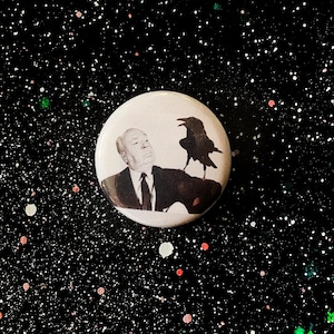 May include: A round, white button featuring a black and white image of a man in a suit with a crow perched on his shoulder. The background is black with white, pink, and green speckles.
