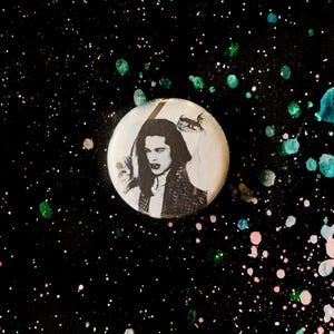 May include: A round, white button with a black and white image of a person holding a sword. The button has a black and white illustration of a small animal. The background is black with colorful paint splatters.