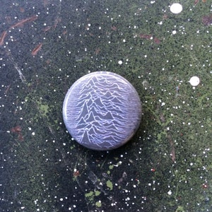 Könnte beinhalten: Ein runder Knopf mit einer schwarz-weißen Grafik eines sich wiederholenden Wellenmusters. Der Knopf befindet sich auf einer grün-weiß gesprenkelten Oberfläche.