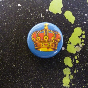 Pode incluir: Um botão redondo com fundo azul apresentando uma ilustração detalhada de uma coroa dourada e vermelha. O botão tem um acabamento brilhante e é provavelmente um alfinete ou distintivo.