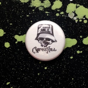 Puede incluir: Un botón blanco con una imagen en blanco y negro de una calavera con un sombrero de pescador con el texto "Cypress Hill" en el sombrero y "Cypress Hill" escrito debajo de la calavera.
