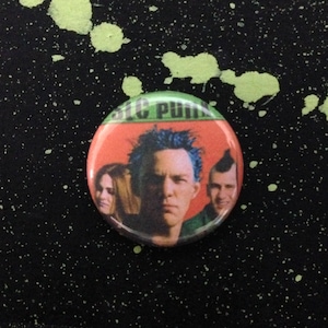Op de afbeelding: Een groene, rode en witte knop met een afbeelding van drie personen. De tekst "Green Day" is zichtbaar op de knop.