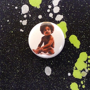 May include: A white button with a black and white image of a young child sitting with their legs crossed. The child is wearing a dark afro and has a light brown complexion.