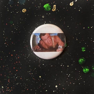 Puede incluir: Un botón blanco con una imagen en blanco y negro de un hombre con la nariz ensangrentada y una expresión de sorpresa. El hombre lleva un tanque blanco y está acostado boca abajo. El botón está sobre un fondo negro con pequeños puntos de colores.