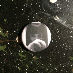 Pode incluir: Um botão preto e branco com um close-up de um rosto com os olhos fechados e as mãos cobrindo a boca.