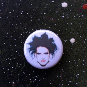 May include: A white button with a black and white image of a person with dark hair and a serious expression.