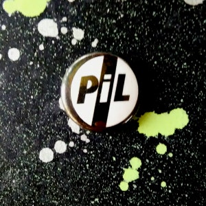 Op de afbeelding: Een witte ovale knop met een zwarte diagonale lijn en de tekst "PiL" in zwarte letters.