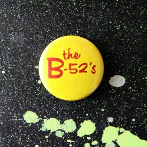 Puede incluir: Un botón amarillo con el texto "the B-52's" en letras rojas.