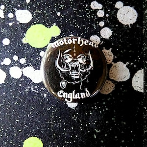 May include: A black and white button with the band name "Motörhead" and the words "England" printed on it. The button features a stylized image of a snarling warthog head with horns.
