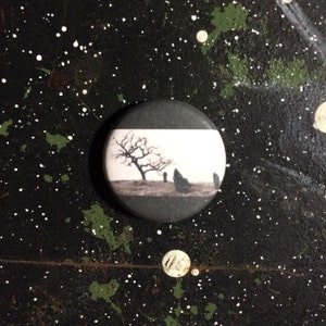 May include: A black and white button with a black border. The button features a black and white image of a tree with bare branches and three figures standing in a field. The background is a white field with a black stripe across the top.