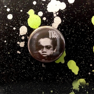 May include: A black and white button featuring a portrait of a person with the word "nas" printed in white on the button.