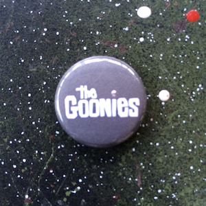 Könnte beinhalten: Ein grauer Knopf mit dem Text "The Goonies" in weißen Buchstaben.