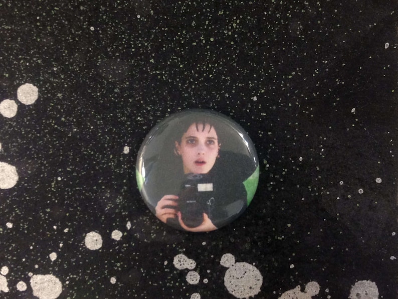 May include: A black and white button featuring a portrait of a young person with dark hair and a camera. The person is wearing a black shirt and has a serious expression.