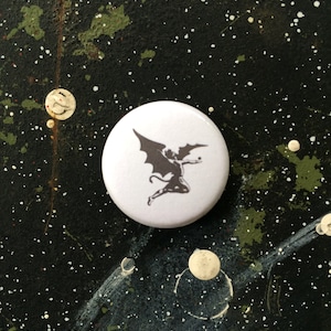 May include: A white button with a black silhouette of a winged demon with a tail.