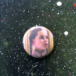 May include: A button featuring a close-up portrait of a young girl with long brown hair in pigtails, wearing a light pink and white collared shirt. The background is a blurry image of a room with a window.