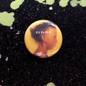 May include: A round, yellow button with the word "GUMMO" in white text. The button features a side profile of a person with dark hair. The background is black with green and white paint splatters.