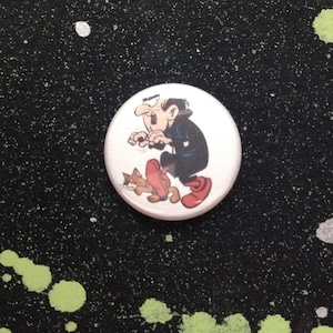 May include: A white button with a cartoon image of a man with a mustache and a cat. The man is wearing a black jacket and red shoes. The cat is brown and white.