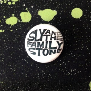Pode incluir: Um botão branco com texto preto que diz "Sly and the Family Stone".