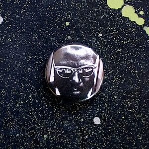 May include: A black and white button featuring a close-up portrait of a man wearing glasses.