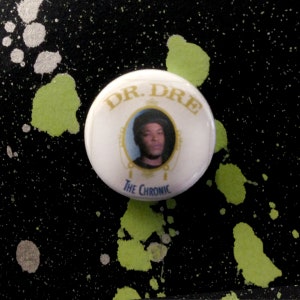 May include: White button with a picture of Dr. Dre on it. The button says "Dr. Dre The Chronic" in gold lettering.