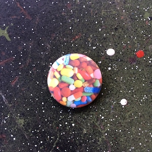 Puede incluir: Un botón redondo que presenta una colorida variedad de pastillas y cápsulas de varias formas y tamaños. El diseño del botón incluye pastillas rojas, amarillas, verdes, azules y rosas, creando un patrón vibrante y llamativo. El botón está sobre un fondo oscuro y moteado.