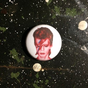 Könnte beinhalten: Ein weißer Button mit einem Porträt einer Person mit roten Haaren und einem roten und blauen Blitz auf dem Gesicht. Der Button befindet sich vor einem dunklen Hintergrund mit weißen und grünen Farbspritzern.