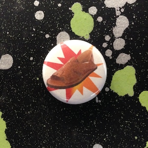 May include: A brown leather sandal with a red and orange starburst design on a white background. The sandal is on a button.