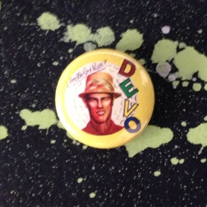 Puede incluir: Botón amarillo con un logotipo DEVO colorido y una imagen de dibujos animados de un hombre con un sombrero. El texto "Are We Not Men?" está escrito en cursiva sobre la cabeza del hombre.