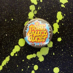 May include: A round button with a black background and a yellow, red, and blue graphic that says "Tribe Called Quest" with a star above the word "Tribe".