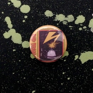 May include: A round button with a yellow lightning bolt and a black and white image of a building with a dome. The button has a yellow and brown border with the text "Bad Brains" printed on it.