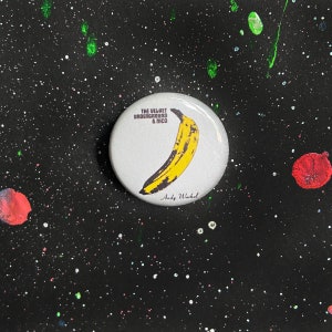 以下が含まれることがあります： 黄色いバナナと「THE VELVET UNDERGROUND & NICO」と「Andy Warhol」の文字が印刷された白いボタンです。