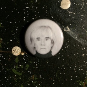 May include: A black and white button featuring a portrait of Andy Warhol. The button is round and has a white background.