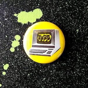 May include: A yellow button with a black and white image of a computer with four faces on the screen. The computer has a keyboard and a monitor.