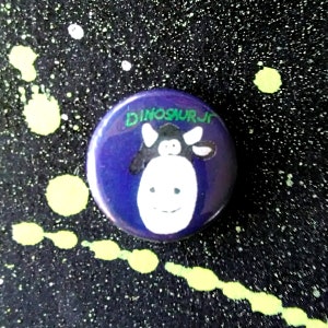 Może przedstawiać: Fioletowy guzik z białą krową w czarne plamy i napisem "Dinosaur Jr." w kolorze zielonym.