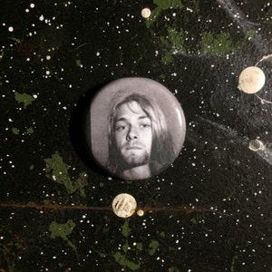 May include: A black and white button featuring a portrait of a man with long hair. The man is looking directly at the camera with a serious expression.