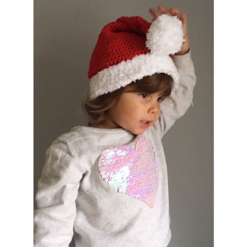 May include: A child wearing a red and white crocheted Santa hat with a white pom-pom on top. The child is wearing a white sweater with a pink iridescent heart-shaped sequin patch.