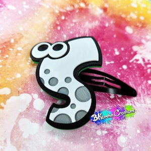 May include: A white and black hair clip in the shape of the number 8 with gray polka dots. The clip has a green border and a black barrette. The background is a colorful mix of pink, orange, and white.