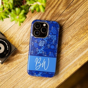 May include: A blue phone case with a floral pattern and the initials "BW" in white script. The case features a dark blue background with lighter blue and white floral accents. The case is on a wooden surface.