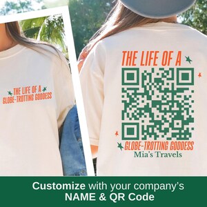 May include: Cream-colored long-sleeve shirt with the text "THE LIFE OF A GLOBE-TROTTING GODDESS" in orange and green. The back of the shirt features a large green QR code and the same text. The shirt is part of a travel-themed collection.