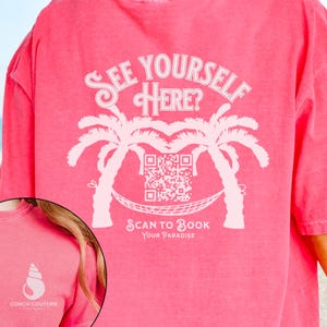 May include: Pink t-shirt with a white graphic of two palm trees with a hammock between them. The graphic also includes a QR code and the text "See Yourself Here? Scan to Book Your Paradise".