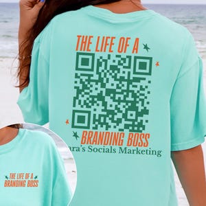 May include: A light teal t-shirt with the text "THE LIFE OF A BRANDING BOSS" in orange and green. A large green QR code is printed on the back. The shirt also has the text "ara's Socials Marketing".