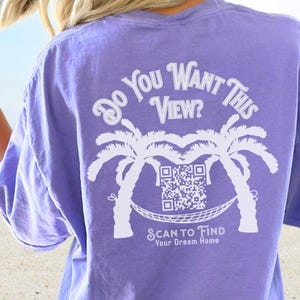 Peut inclure: T-shirt violet avec un motif blanc représentant des palmiers, un hamac et un code QR. Le texte sur le t-shirt est "Do You Want This View? Scan to Find Your Dream Home."