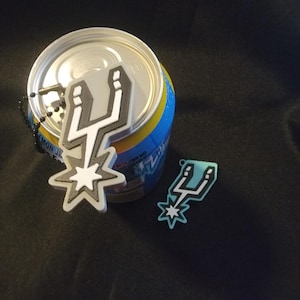May include: Two pendant-style keychains. One is a large, gray and white design with a starburst base, and the other is a smaller, teal and white version. A can of Lemon Juice is in the background.