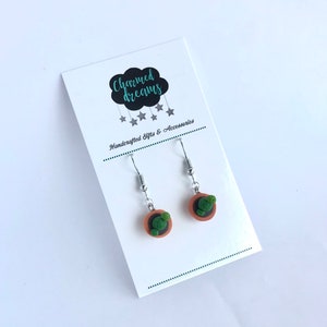 Puede incluir: Un par de pendientes de plata con peque&ntilde;os maceteros de arcilla y cactus verdes. Los pendientes est&aacute;n en una tarjeta blanca con el texto "Charmed Dreams" y "Handcrafted Gifts & Accessories".