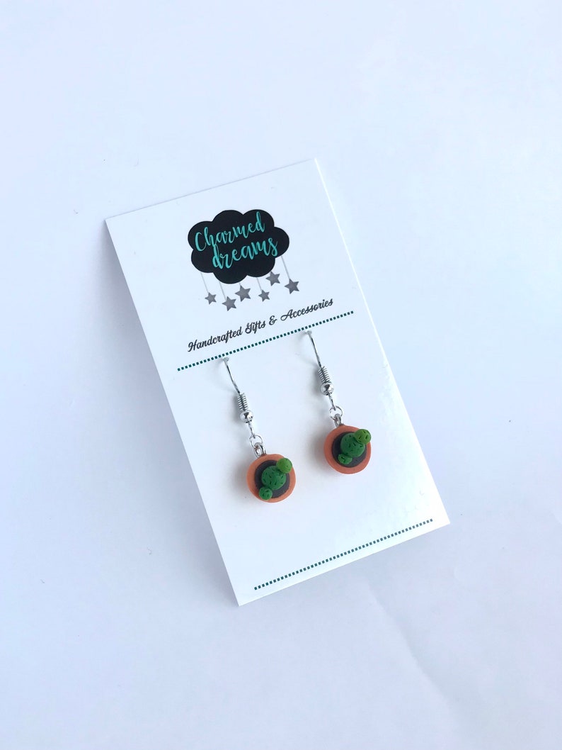 Puede incluir: Un par de pendientes de plata con peque&ntilde;os maceteros de arcilla y cactus verdes. Los pendientes est&aacute;n en una tarjeta blanca con el texto "Charmed Dreams" y "Handcrafted Gifts & Accessories".