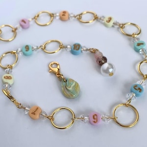 May include: A gold-coloured chain bracelet featuring pastel-coloured number beads, clear crystal beads, and gold-coloured circular links. A teardrop-shaped charm and a pearl bead dangle from the bracelet. The numbers on the beads are from 2 to 10.