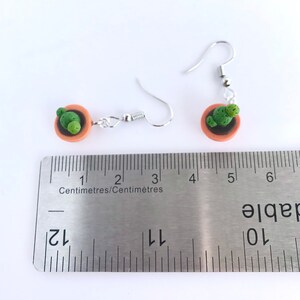 Puede incluir: Un par de pendientes de plata con un peque&ntilde;o tiesto de terracota y un amuleto de cactus verde. Los pendientes miden aproximadamente 3,2 cm de largo.